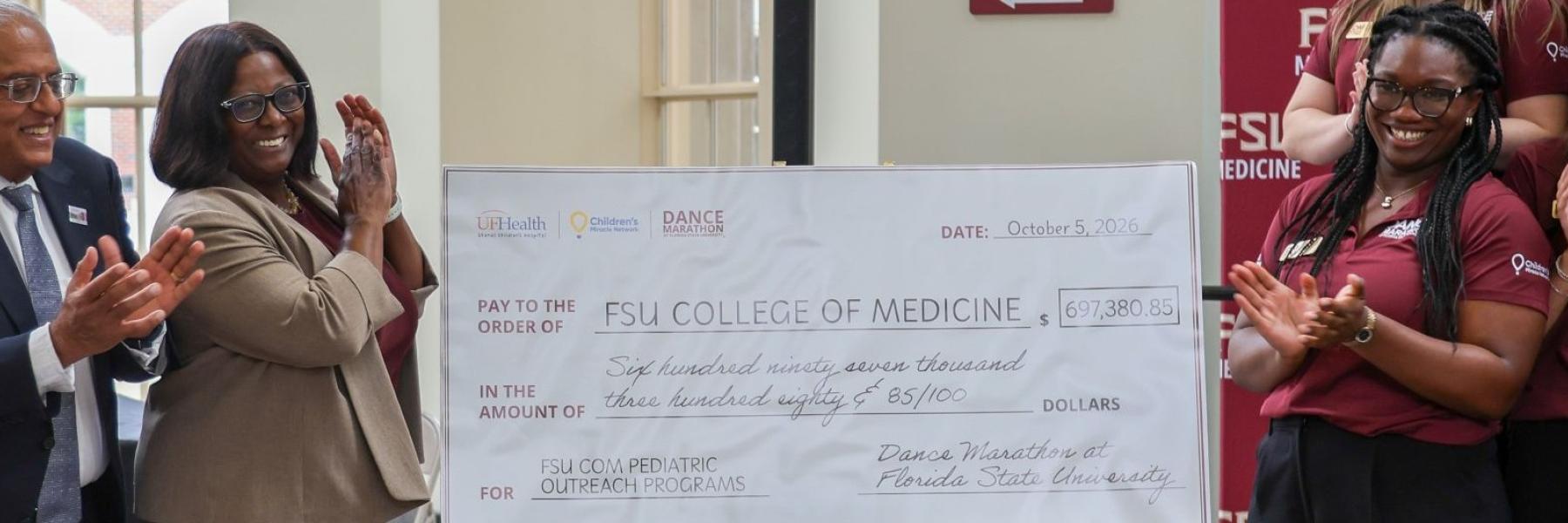 Dr. Savani and Dean Littles stand to the left of Dance Marathon's ceremonial check to FSU College of Medicine, while DM@FSU 2027 Executive Director Emmanuella Essien and the executive board stand to the right of it.
