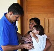 A medical professional in a blue shirt uses a stethoscope to examine a young child held by their mother in a doorway.
