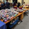 A man and women speak to a group of people standing next to a table of sensory comfort items to be stuffed into bags.