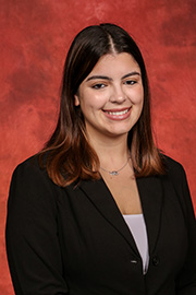 A smiling woman with shoulder-length dark hair and a black blazer stands against a reddish-brown textured background.
