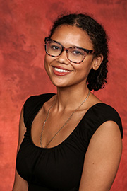 A smiling woman with curly dark hair and glasses wearing a black top stands against a reddish-brown textured background.