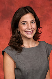 A woman with wavy brown hair and a grey floral top smiles in a studio portrait against a reddish-brown background.