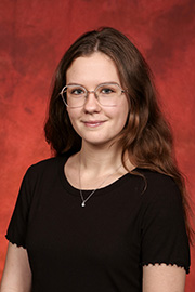 A woman with long wavy brown hair and round glasses wearing a black shirt smiles in a studio portrait against a reddish-brown background.