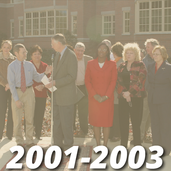 Timeline from 2001-2003, image shows a group of about twelve adults dressed in professional attire standing outside the college of medicine building with one adult male speaker standing with a microphone ready to speak, the other adults are standing behind the speaker as if they are supporting his speech.
