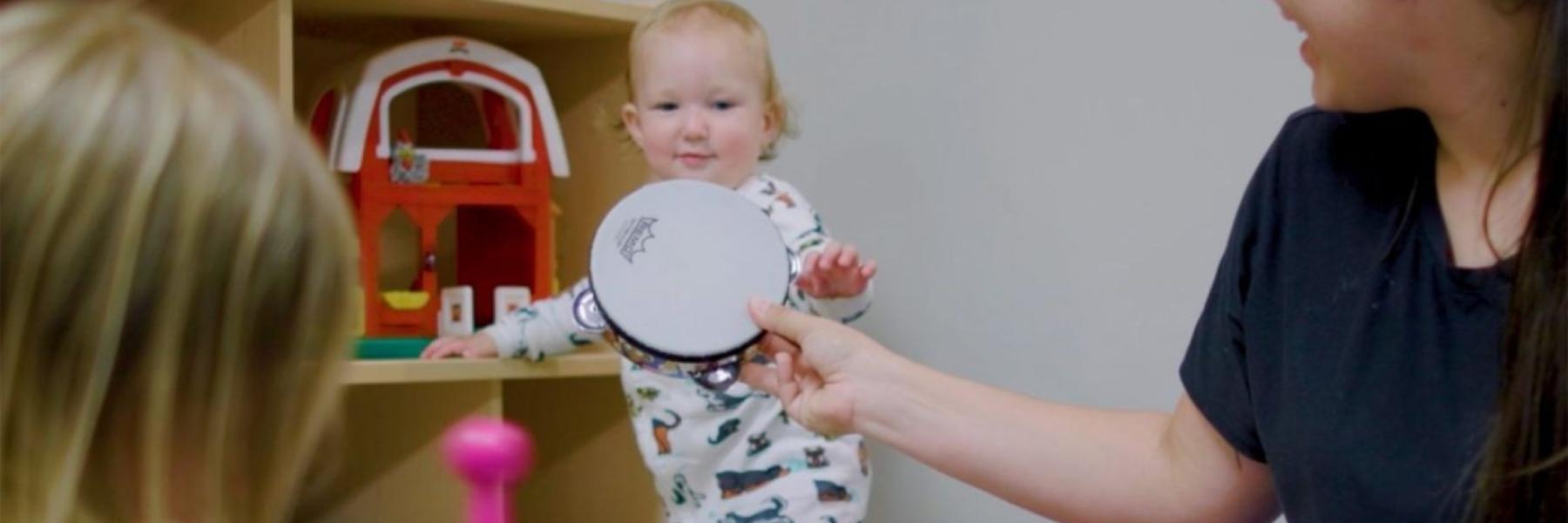 A woman hands a toy tambourine to a toddler.