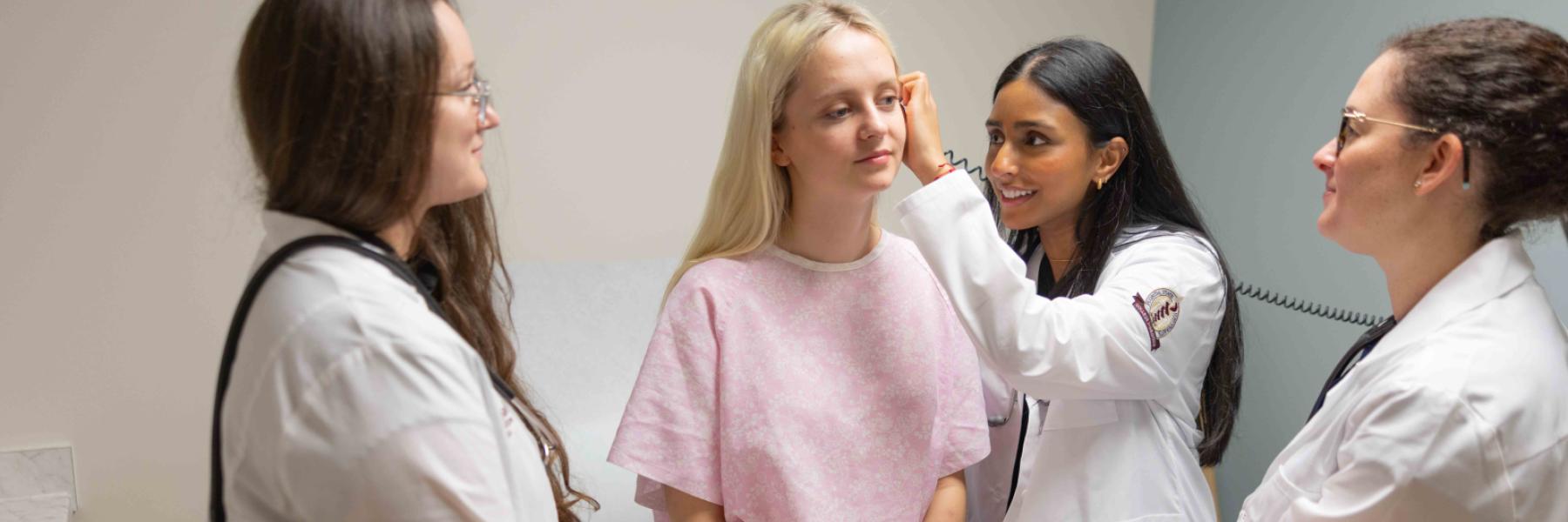 Two medical students in white coats with a blond, young female patient sitting on a bed. One student is looking in the patient's ear, while a physician looks on.